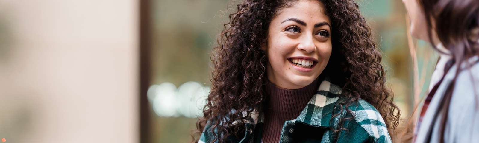 A young woman with long curly hair is smiling warmly while engaged in conversation with another person whose face is not fully visible. She is wearing a green plaid jacket over a maroon ribbed turtleneck sweater. The setting appears to be outdoors in an urban or public space with a softly blurred background. The woman represents a care navigator, offering approachable and supportive communication, possibly in a healthcare or community service context.