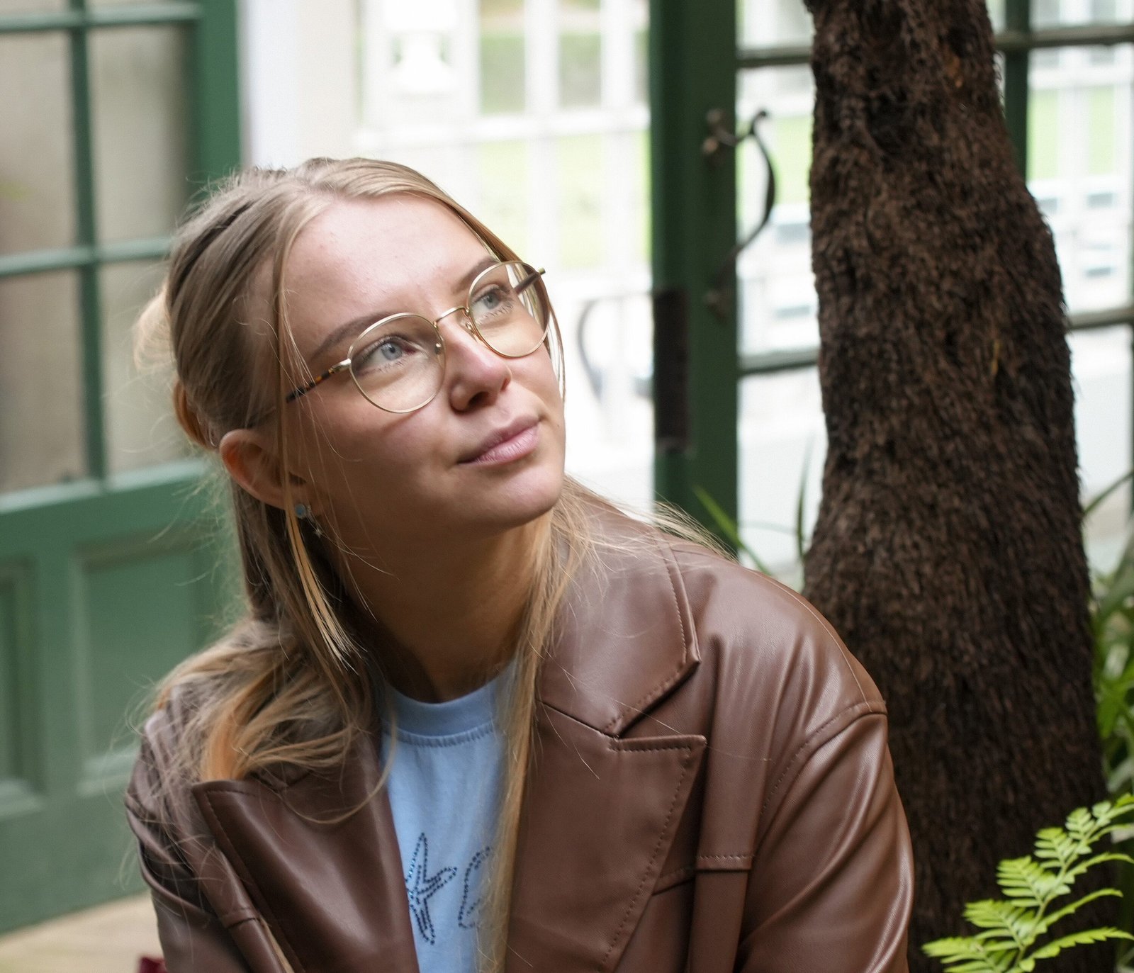 A young woman stands in a park, wearing a brown coat and blue jeans. She has long blonde hair and is smiling softly. The pathway is flanked by benches and lush greenery, with autumn foliage in the background, creating a serene and picturesque atmosphere.